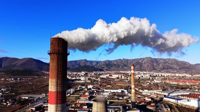 Industrial chimney emitting smoke over city with mountains, Bulgaria. Elevated drone view of tall brick stacks releasing white plume, urban sprawl and factory rooftops in middle distance.