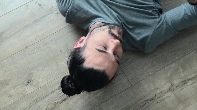 Contemplative man lying on wooden laminate floor with hands behind head removes round glasses, leaves frame after quiet side gaze, overhead home portrait with window reflections and soft natural light
