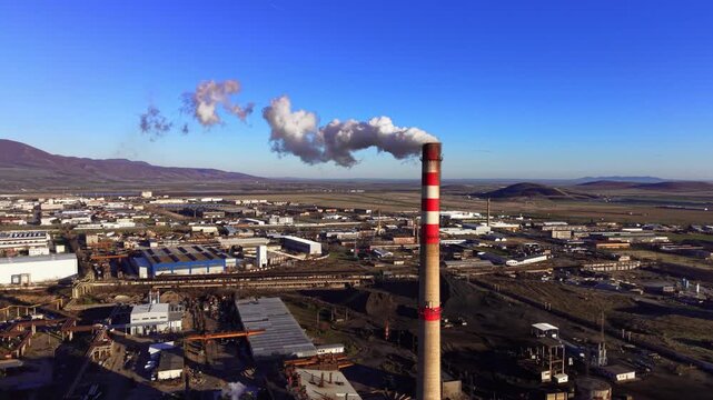 Striped industrial chimney releasing smoke over factory zone, Bulgaria. Drone view of tall stack with smoke plume, industrial plain with warehouses, roads, rail lines.