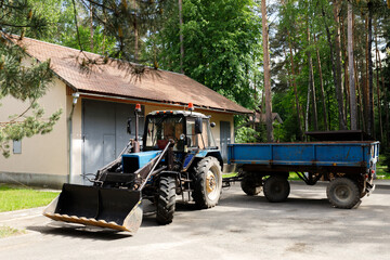 Blue industrial tractor with front loader bucket and trailer parked near garage building. Utility machinery vehicle for agriculture or construction work. Rural countryside lifestyle concept title © Tina