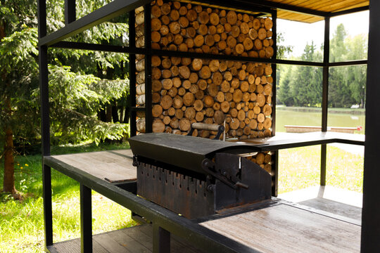 Empty outdoor kitchen pavilion with black metal grill barbecue area. Modern backyard terrace with fireplace and stacked firewood wall decoration near lake. Summer backyard bbq picnic concept