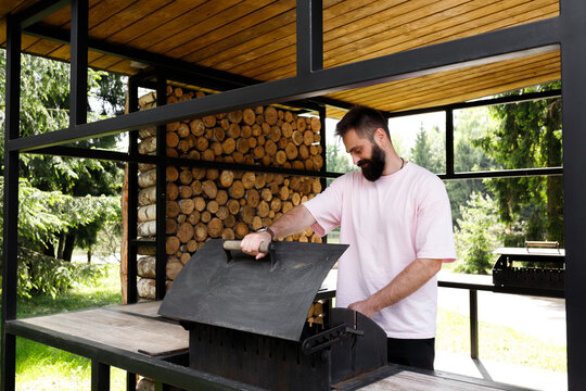 Bearded man opening black metal grill lid at backyard barbecue area. Male chef preparing wood fire bbq inside modern outdoor pavilion kitchen. Firewood background, summer food cooking concept