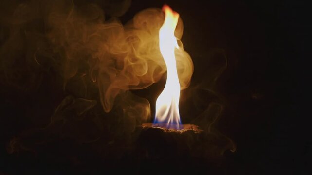 Igniting charred disk producing blue core yellow-orange tip flame in dark studio, with smoke
