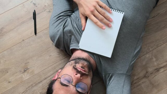 Long haired man in glasses studying handwritten notebook notes while lying on wooden floor, resting with journal on chest, thinking quietly, then getting up and leaving calm slow life home scene