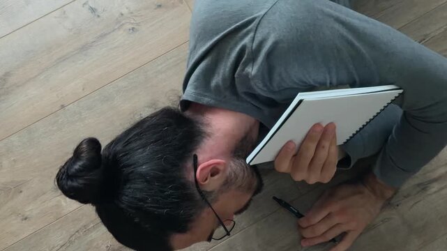 Thoughtful writer in glasses lying on wooden floor reading notebook, then turning over to grab pen and write fresh idea, overhead home interior scene of relaxed creative workflow in soft natural light
