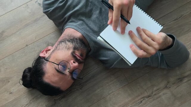 Thoughtful writer lying on floor with notebook and pen, turning pages, reading drafts and studying ideas during relaxed creative writing process at home in natural window light, high angle close up