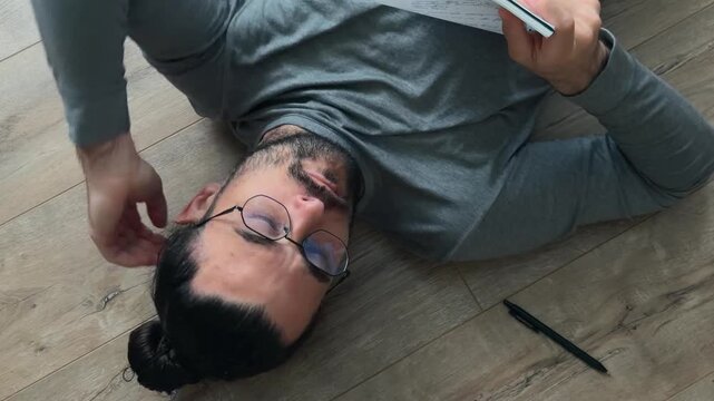 Thoughtful writer in glasses lying on floor with notebook on chest, flipping pages and reading handwritten notes, resting with hand behind head during calm creative planning at home in natural light