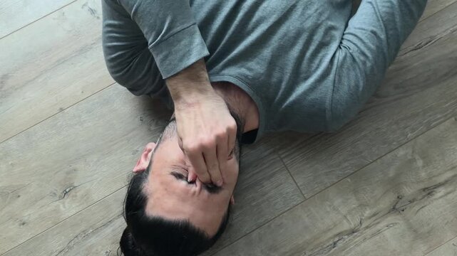 Tired man lying on wooden laminate floor rubs eyes, puts round glasses on, folds both hands behind head and rests in quiet thought, overhead home portrait with soft window glare and natural light