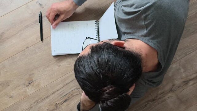 Unrecognizable long haired man in glasses reviewing handwritten notebook notes while lying on side at home, calm slow life planning, private reflection and creative workflow from overhead view