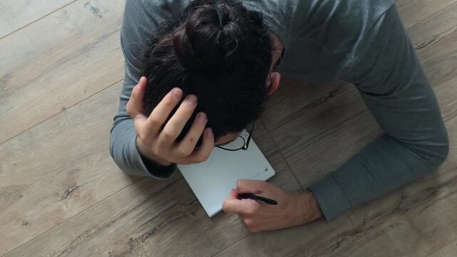 Unrecognizable long haired writer lying on floor with notebook, supporting head by hand while writing notes in creative workflow, overhead view of journaling, planning, focused solitude at home