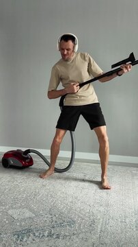 Energetic barefoot man having fun cleaning the house. He is listening to music on headphones and pretending to play a guitar with a vacuum cleaner