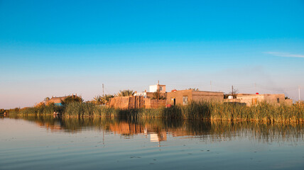 Obraz premium Traditional houses reflect in tranquil waters of Iraqs Marshes at sunset