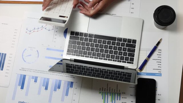 Top-down view of a male accountant analyzing financial charts and typing on a laptop keyboard, using a calculator for budget calculations and reviewing business project reports