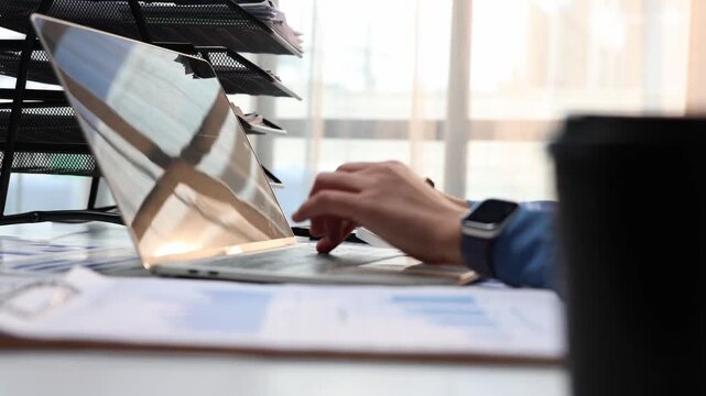 Financial analyst typing on a laptop and checking business data on documents with graphs and charts while sitting at a modern office desk with a cup of coffee nearby