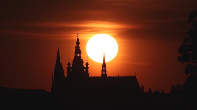 Golden sun sets behind historic spires of Prague Castle. Silhouetted towers stand stark against the glow. Warm hues wash over the horizon. Dramatic light frames historic architecture of Bohemia, Czech