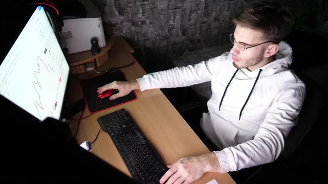 Young guy working on computer at evening. Male broker analyzing real-time commodities and foreign exchange graphs. Financial analyst trading online on stock market at night. Concept of freelance job