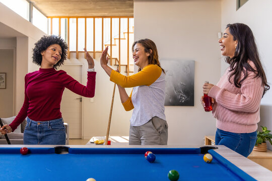 Diverse female friends high-fiving around blue felt pool table in home, holding cues and red bottle