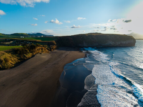 Aerial view of the dark volcanic sands of a secluded beach bordered by steep cliffs and lush green hills under a bright blue sky Sao Miguel, Azores, Portugal.