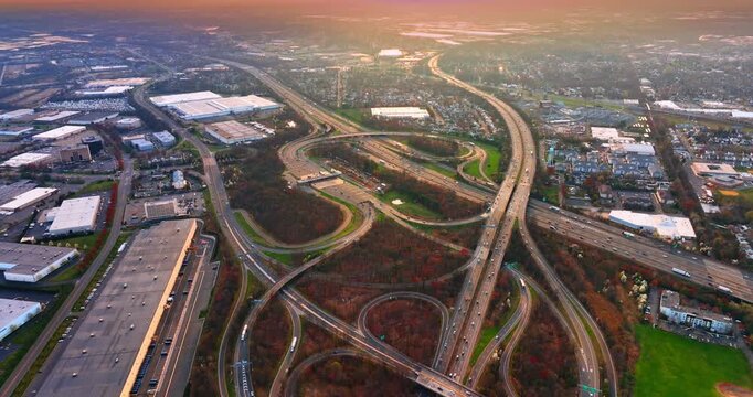 Sprawling freeway junction through suburban landscape at dusk, New Jersey. Looping ramps and warehouse rooftops spread toward a glowing horizon with autumn-tinged trees lining the roads