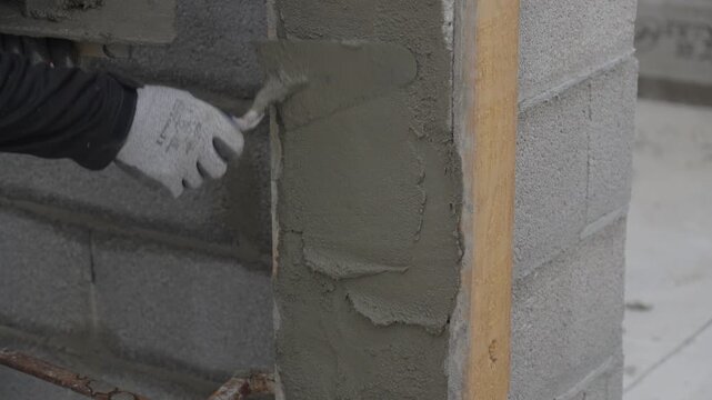 Construction worker applying fresh cement mortar to a cinder block wall with a trowel. Professional plastering and masonry work on a building site.
