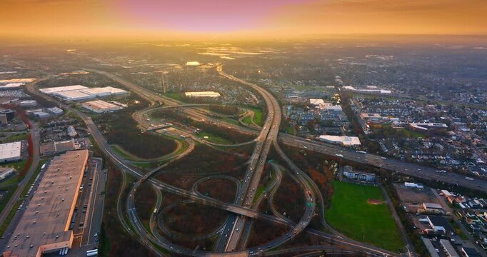 Aerial panorama of a freeway interchange at golden sunset, New Jersey. Wide elevated view of looping ramps and warehouses spreading toward a hazy glowing horizon beneath orange sky