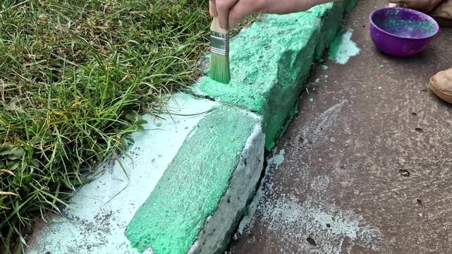person squatting down and using brush paint concrete curb green to blend in with the adjacent lawn. activity is often subject of joke meme. It usually refers to an absurd pursuit of perfection.