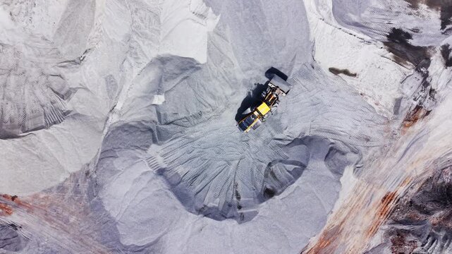 Drone view of yellow heavy machinery working on large textured mounds of grey gravel. Overhead shot of wheel loader moving sand aggregate pile