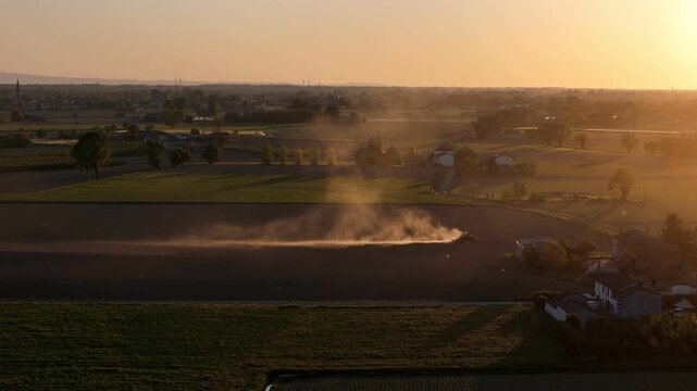 Tractor performs harrowing and seedbed preparation during May spring cultivation in Province of Piacenza Emilia Romagna Italy creating illuminated dust cloud at sunset, slow motion aerial slide