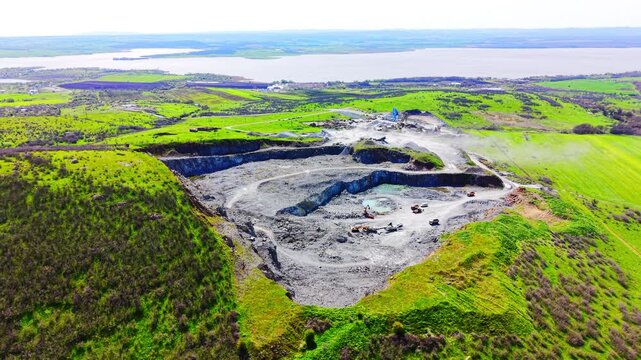 Panoramic drone shot of stone excavation crater surrounded by green hills with water body in background. Wide aerial view of open pit mine near coastal lake, Bulgaria.