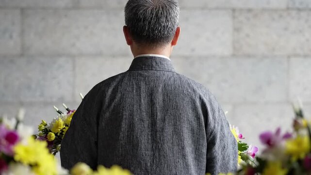 Man in traditional grey jacket stands before large floral arrangement of yellow and white chrysanthemums and pink orchids, formal occasion