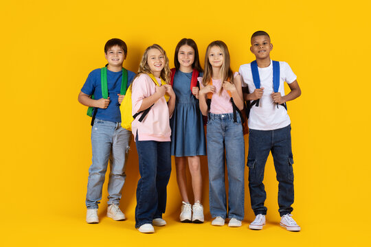 Diverse group of smiling school children with backpacks posing together on bright yellow studio background, representing friendship, education, teamwork and back to school concept