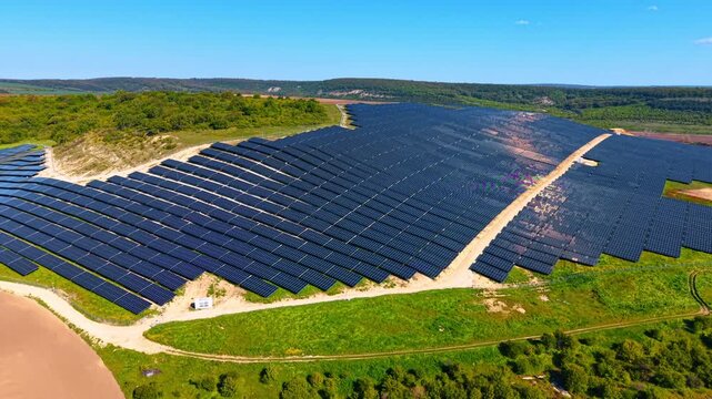 Aerial view of solar plant surrounded by green countryside. Drone angle on angled panel ranks ringed by meadow, scrubland and forested ridges in the distance.