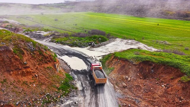 Elevated view of loaded commercial vehicle on a muddy track heading towards bright green rural fields. Red tipper truck driving out of quarry site.