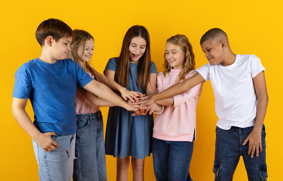 School children stacking hands together on yellow studio background, symbolizing teamwork, unity, cooperation and friendship in education environment