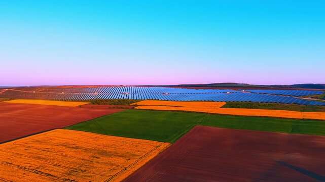 Solar plant at twilight beyond colorful patchwork crop fields. Aerial over a mosaic of canola, green and plowed plots leading to module rows under a pastel sky.