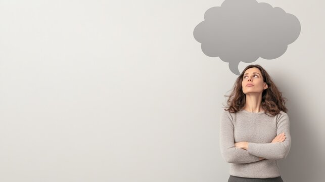 Pensive businesswoman is thinking with arms crossed and looking up at an empty gray speech bubble above her head, suggesting communication, questioning, and messaging concepts