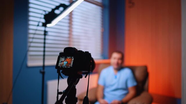 Rear screen shows seated presenter in warm consultation studio. Camera display previews a man in scrubs in an armchair under orange and blue accent lighting.