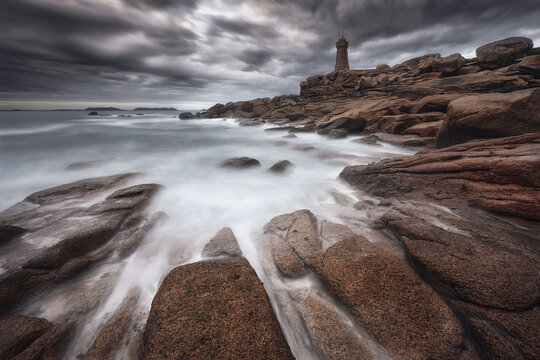 View of the Phare de Ploumanac'h lighthouse standing on the pink granite coast with misty long exposure waves under a dramatic cloudy sky in Brittany, France.