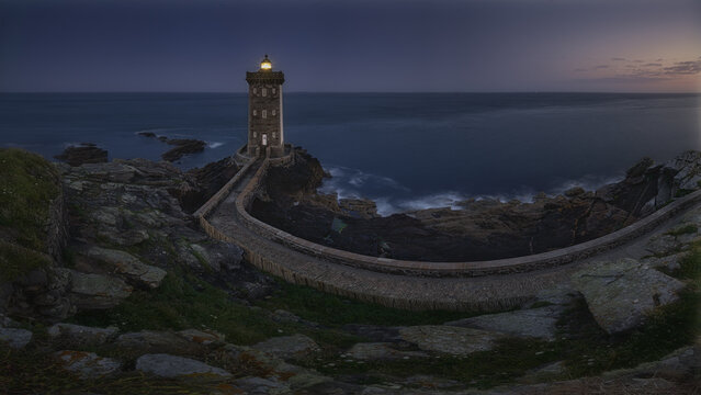 View of the Phare du Petit Minou lighthouse at dusk with its illuminated lantern and a curved stone bridge along the rocky coastline of Brittany, France.