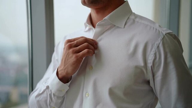 Adjusting man in white dress shirt smoothing collar and front by tall window, preparing for meeting