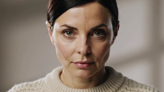 Recording starting, woman in cream knit sweater opening eyes and holding gaze in neutral room