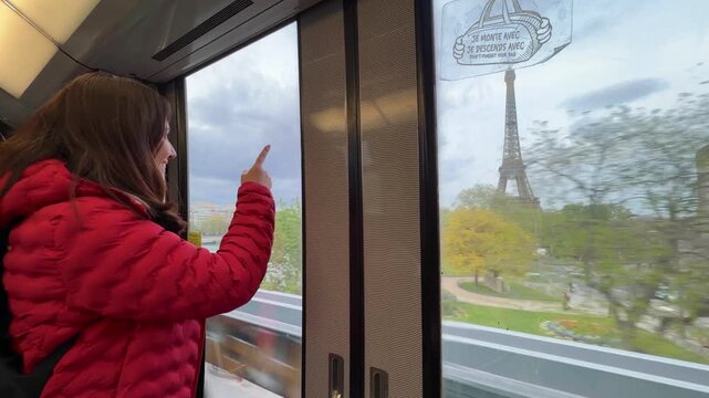 Young tourist woman in a red jacket excitedly pointing at the Eiffel Tower while traveling on an elevated metro line, enjoying the scenic view of Paris and the Seine river during her vacation