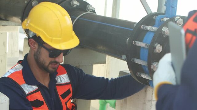 Engineers with safety vests and helmets maintain a cooling tower on rooftop of solar farm. Two workers inspect cooling system of building. Male technician with coworker discusses during work together.