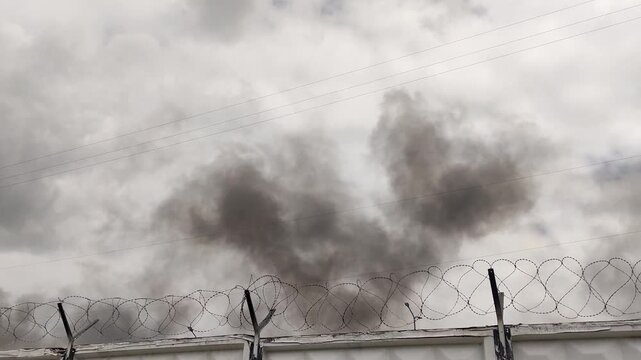 Thick black smoke rising in the sky above a white concrete wall with barbed wire