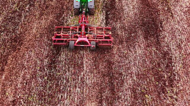 Red cultivator frame in full detail with tines in soil, green tractor cab visible at top, red-brown textured ground fills frame. Overhead detail of red disc harrow attachment tilling soil