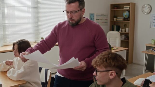 Male teacher walking among desks with focused students and giving out test papers for final exams in modern classroom