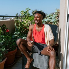 Fototapeta premium Young man relaxing on a sunny balcony with coffee among potted plants for lifestyle background and social media banner design