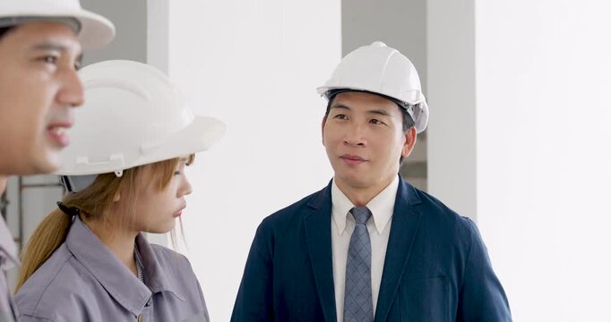 Asian male inspector talks with female engineer and site worker, pointing across unfinished building as construction quality control inspection and progress meeting continues indoors today daily.