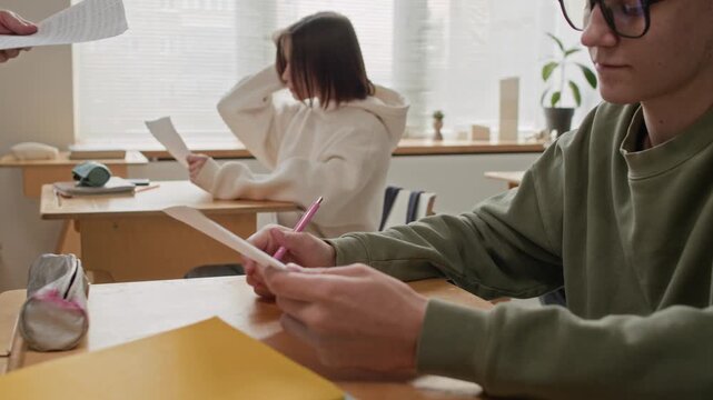 Low angle shot of teenage boy with curly hair and large glasses receiving test paper while sitting at his desk among other students in modern classroom