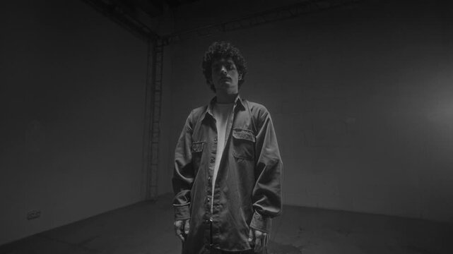 Cinematic black and white shot of Caucasian young man standing in dark studio illuminated by flashing lights and looking at camera intencely, accentuated by laght and shadows, copy space